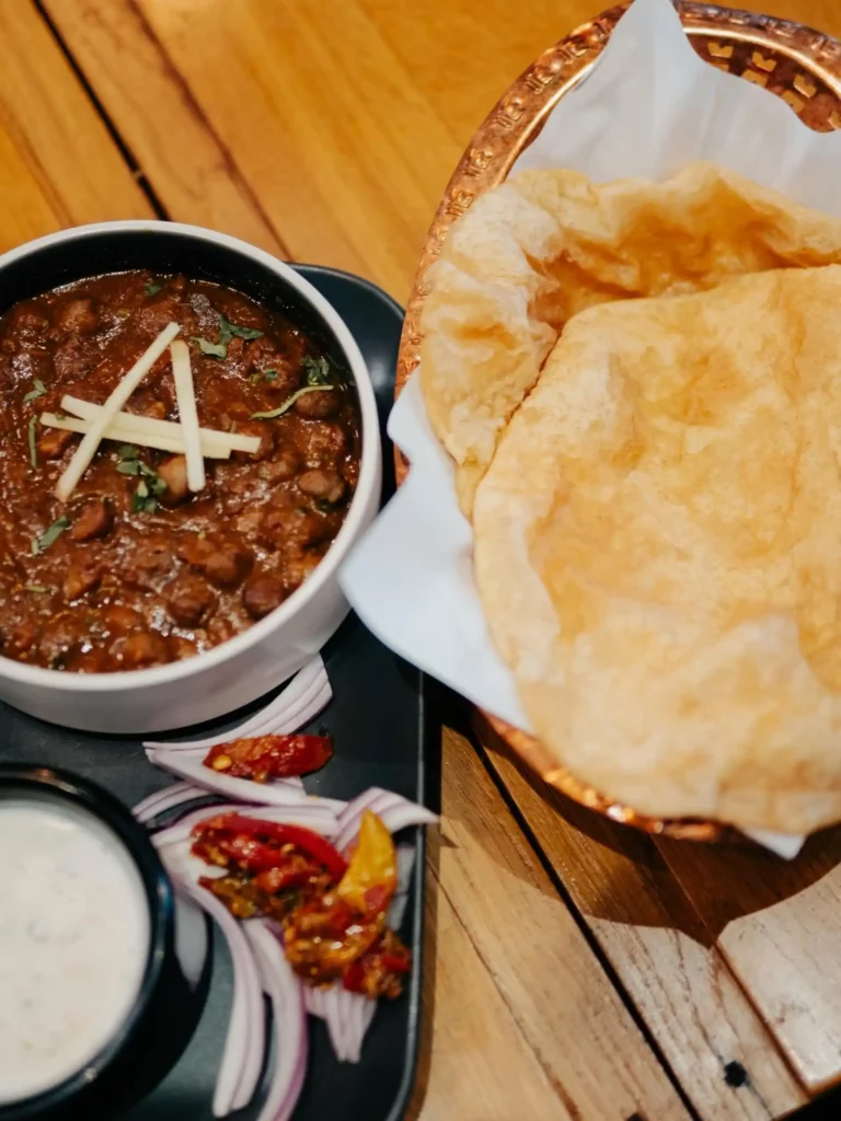 Channa Puri served with fluffy deep-fried puri bread, spicy chickpea curry, raita, pickled vegetables, and fresh onions.